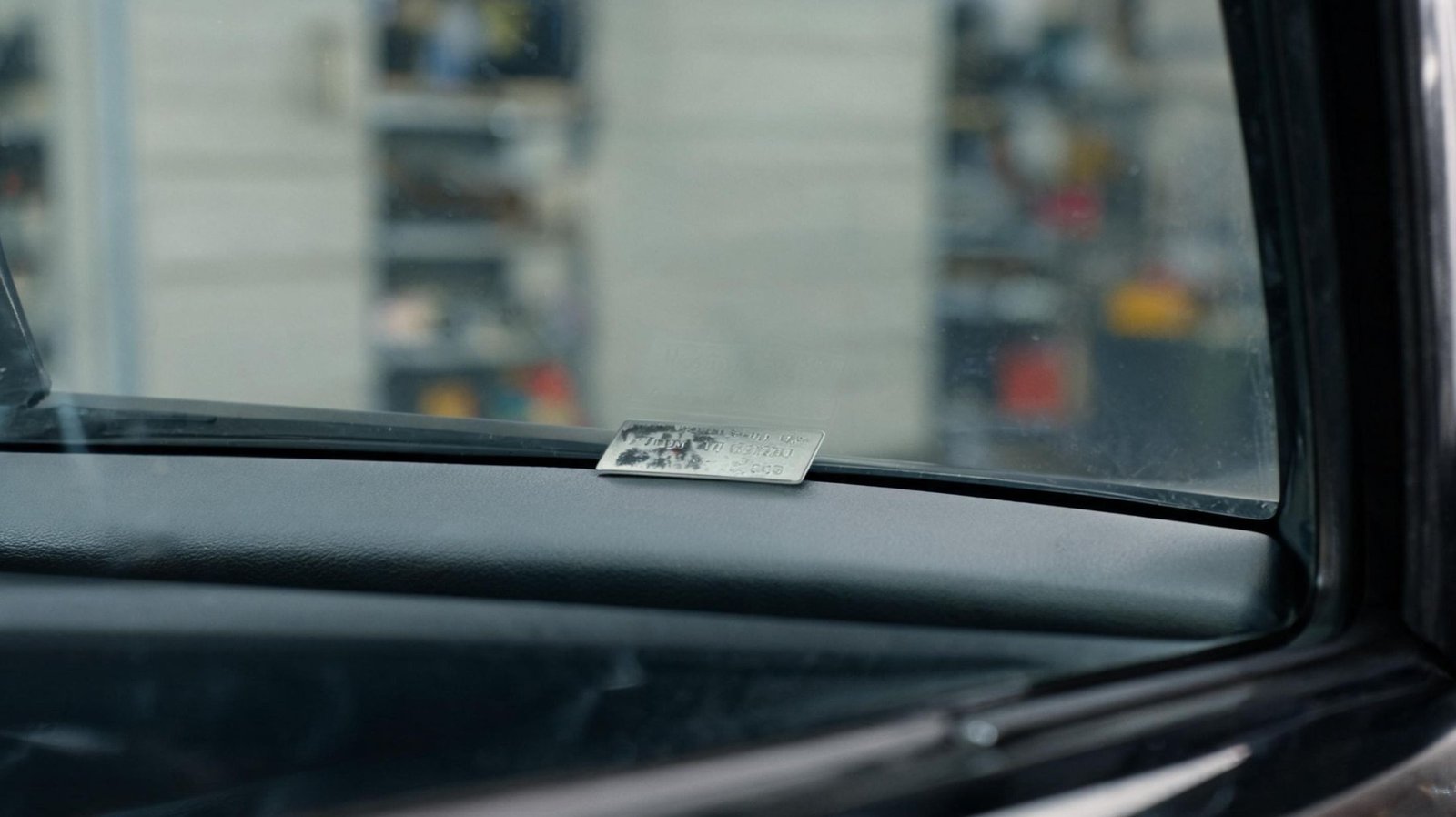 Small metal VIN plate on a dashboard edge photographed through a windshield from outside, with blurred illegible characters and a garage in the background